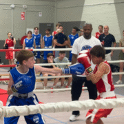 Actiefoto van twee jeugdige boksers van Boxing University Breda in de ring tijdens een sparringsessie voor het boksen in Breda. De bokser in het blauw reikt met een stoot naar zijn tegenstander in het rood. Op de achtergrond kijken toeschouwers en andere boksers toe.