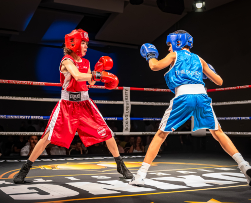 Twee jeugdige boksers, één in rood en één in blauw, staan tegenover elkaar in een boksring tijdens een amateurbokswedstrijd op het Boxing University Breda boksgala. Het publiek kijkt toe vanaf de tribune.